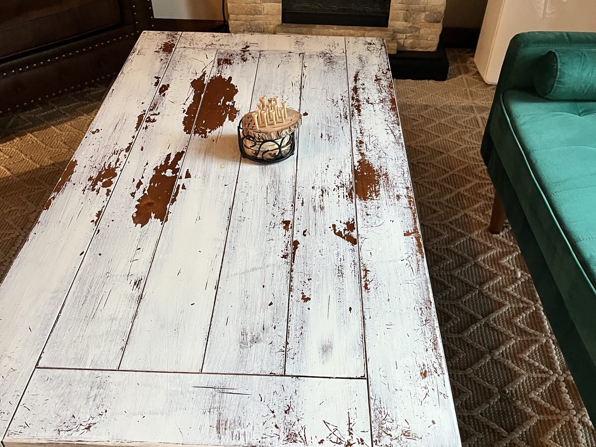White distressed plank-top coffee table with shaped dark metal legs in front of a stone fireplace